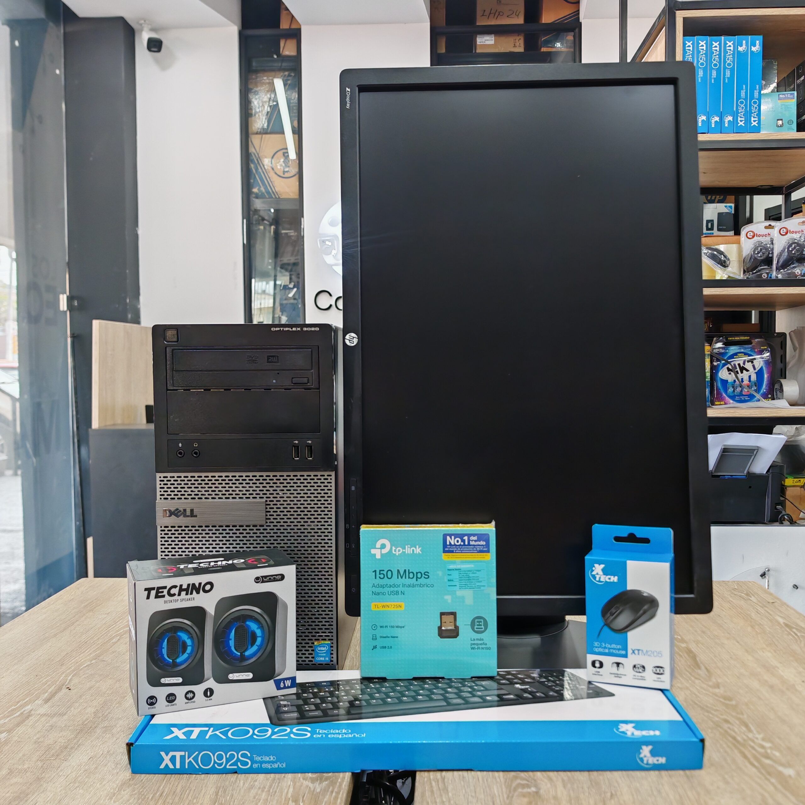 Fotografía de una computadora de escritorio Dell Optiplex junto a un monitor de 22 pulgadas que muestra el precio de Q2,665. En frente se ven las cajas de los accesorios incluidos: bocinas Techno, adaptador Wi-Fi TP-Link y mouse Xtech. El fondo muestra el interior de la tienda Compusdeguate con la dirección y teléfono en la parte inferior.